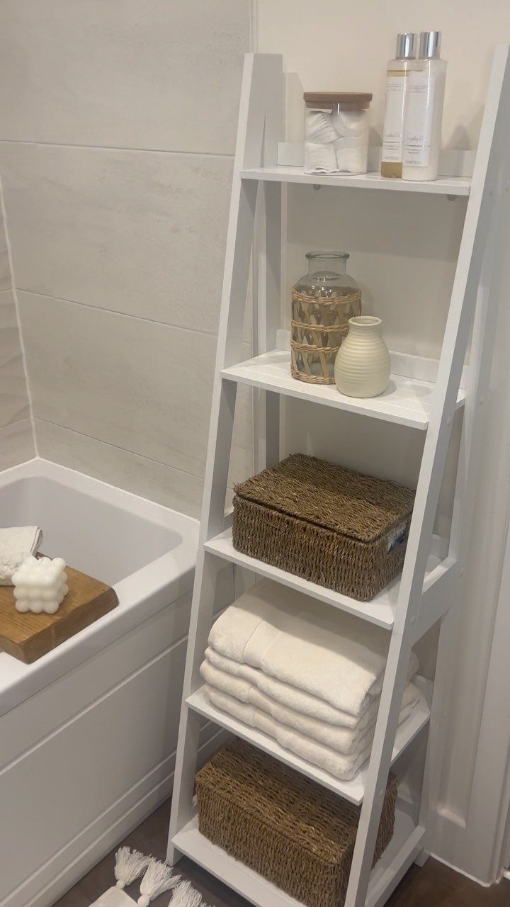 14 Bathroom Small Space Storage Ideas That Actually Fit 4 White ladder shelf holding towels, wicker baskets, and bathroom accessories next to a white bathtub.
