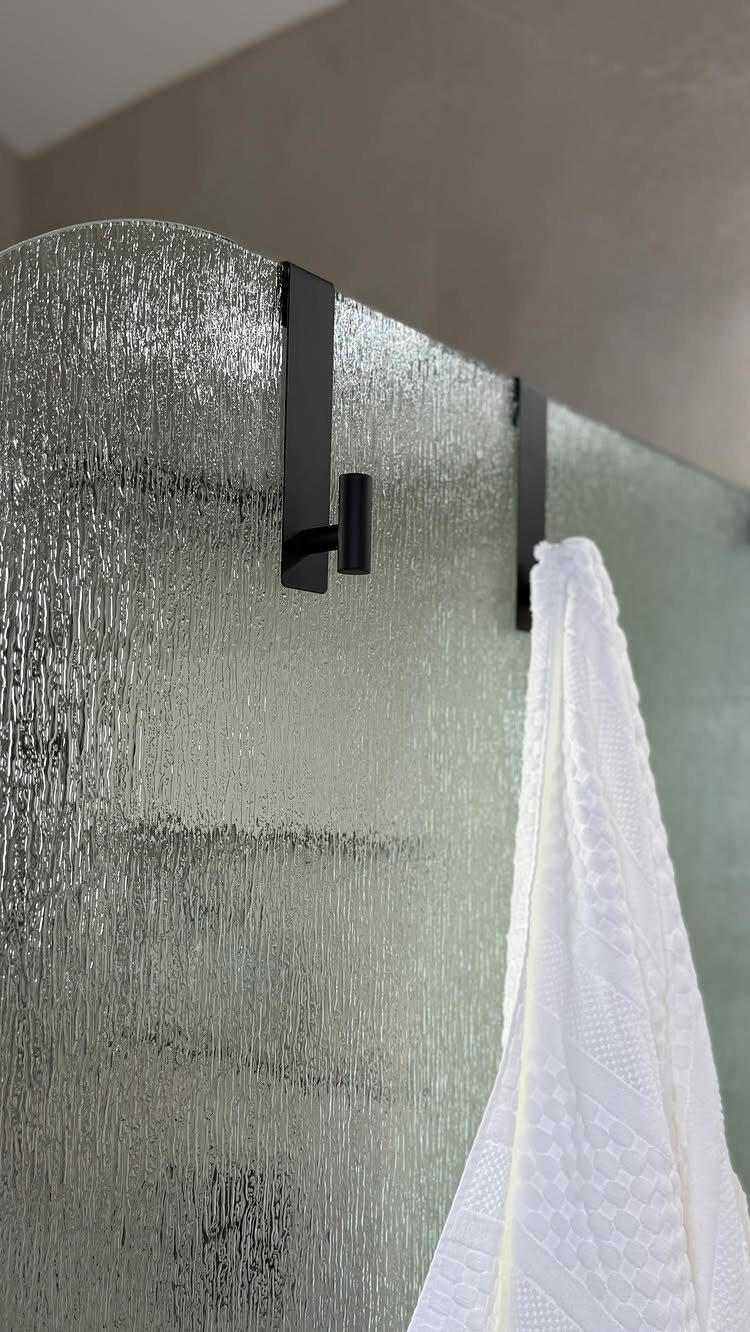14 Bathroom Small Space Storage Ideas That Actually Fit 11 Black metal towel hooks mounted on a textured glass shower door with a white towel.