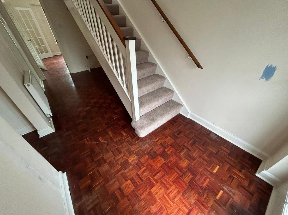 Reddish-brown parquet wood flooring in a hallway next to a carpeted staircase with white railings.