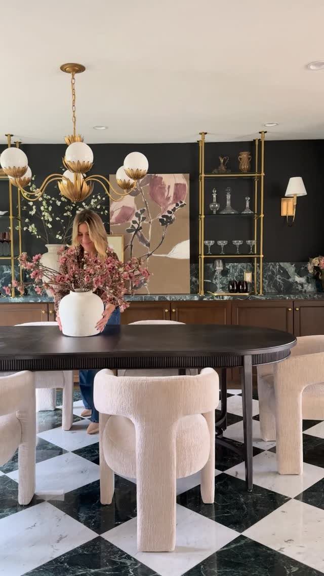 A person arranges pink flowers in a white vase on a dark dining table.