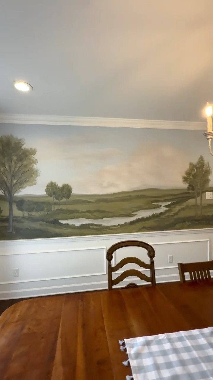 Landscape mural of green fields and trees painted on a dining room wall above wainscoting.