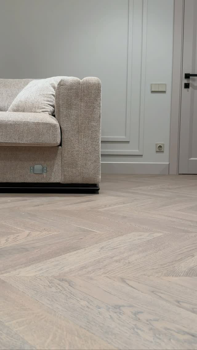 Light grey wood floors in a herringbone pattern next to a beige sofa and white wall.