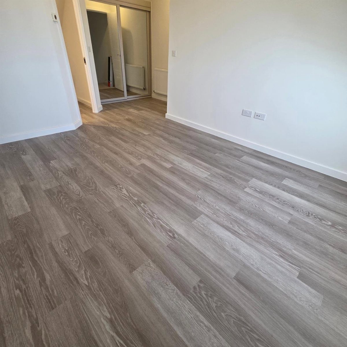 Grey wood-effect laminate flooring planks covering the floor of an empty room with white walls.