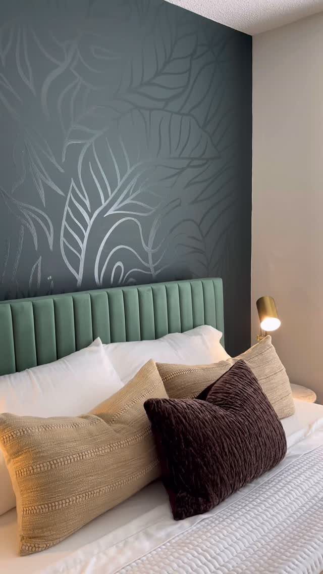 Dark teal accent wall with large leaf patterns behind a green velvet headboard and bed.