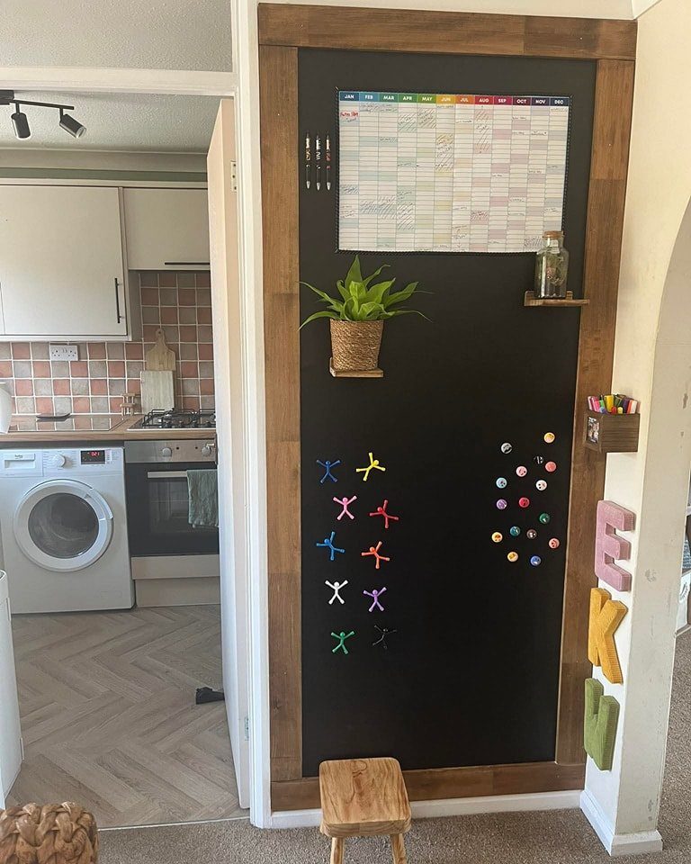 Black chalkboard wall with magnets, a calendar, and a small plant in a wooden frame.