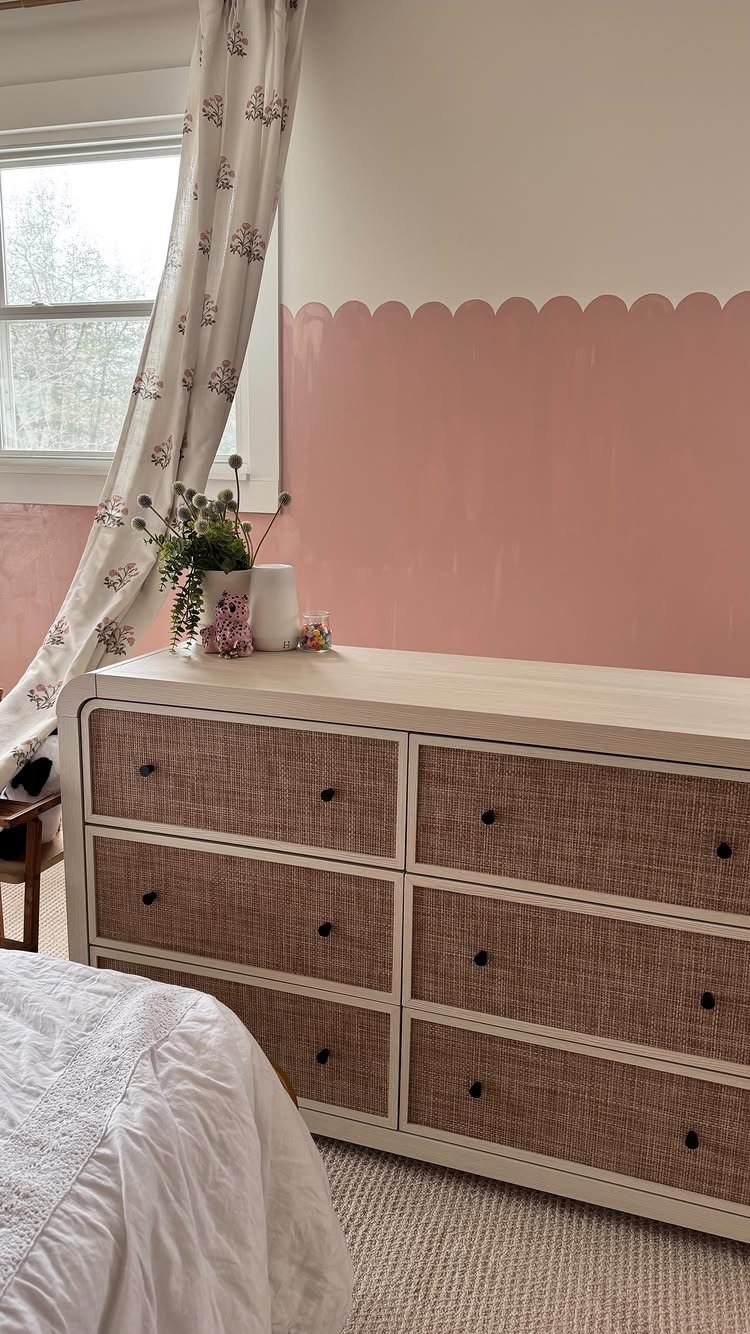 A pink scalloped paint design on a white wall behind a light wood dresser.