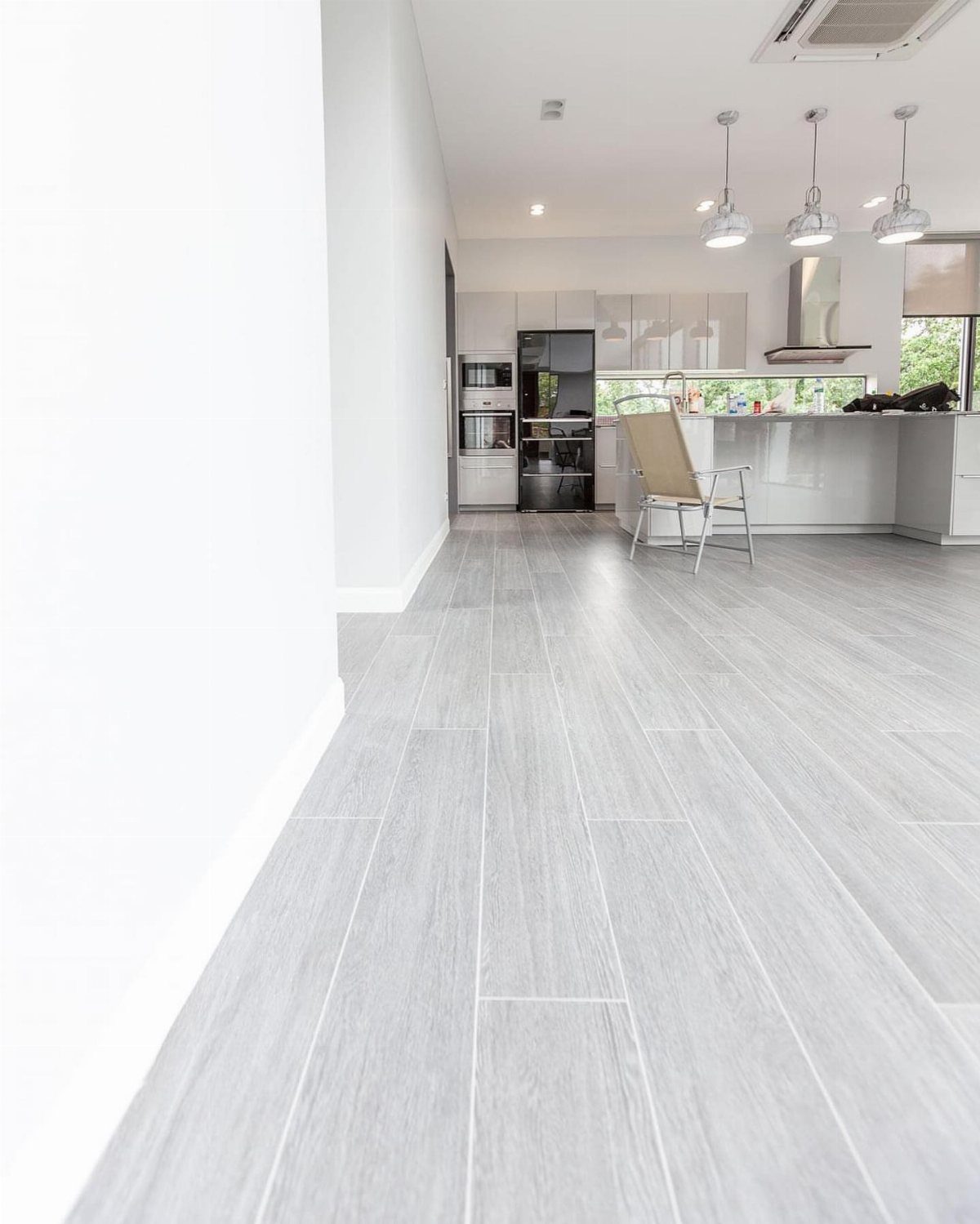 11 Kitchen Flooring Trends for 2026 That Actually Hold Up 3 Light gray wood-look porcelain tiles covering the floor of a modern, bright kitchen.