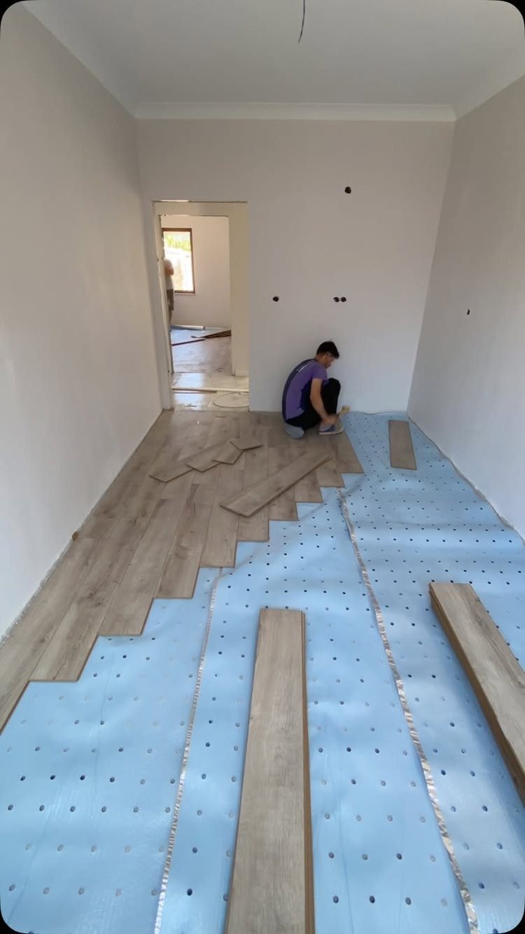 A person installs light oak wood-look planks over blue underlayment in a room.