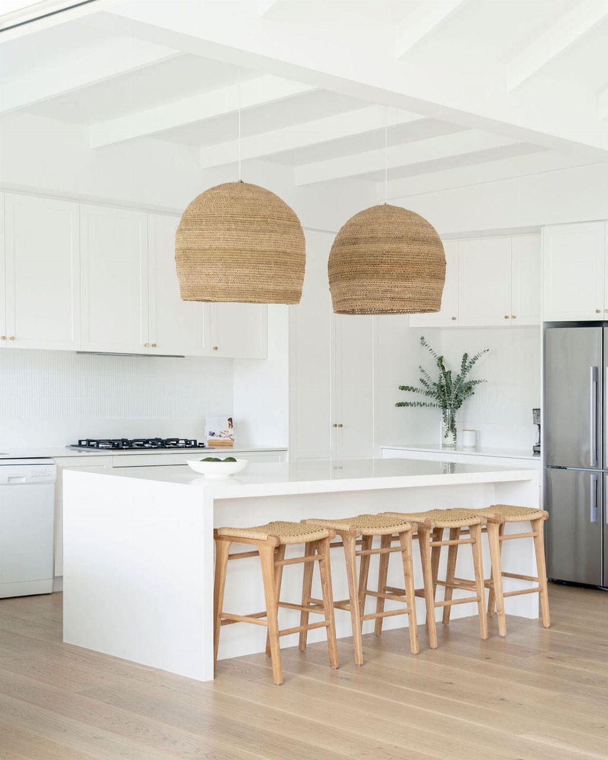 Four wooden backless stools tucked under a white kitchen island with woven pendant lights above.