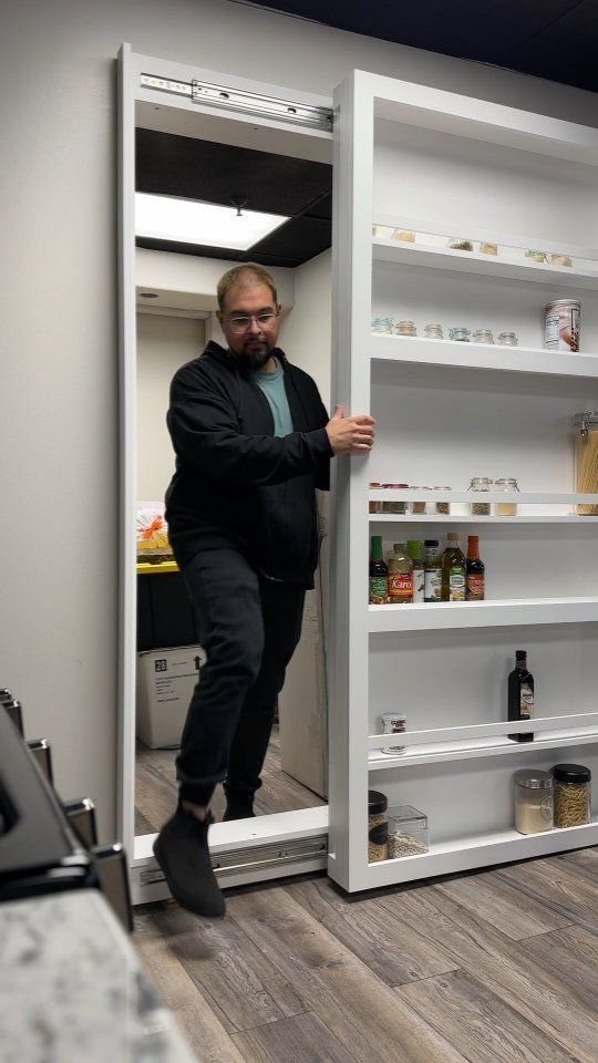 A man opening a white sliding door pantry system filled with organized shelves of jars and bottles.