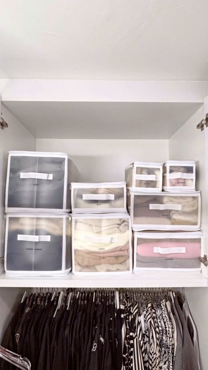 8 Storage Closet Organization Ideas for Tighter Spaces 2026 7 Clear plastic storage bins filled with folded clothes sit on a white closet shelf above hanging garments.