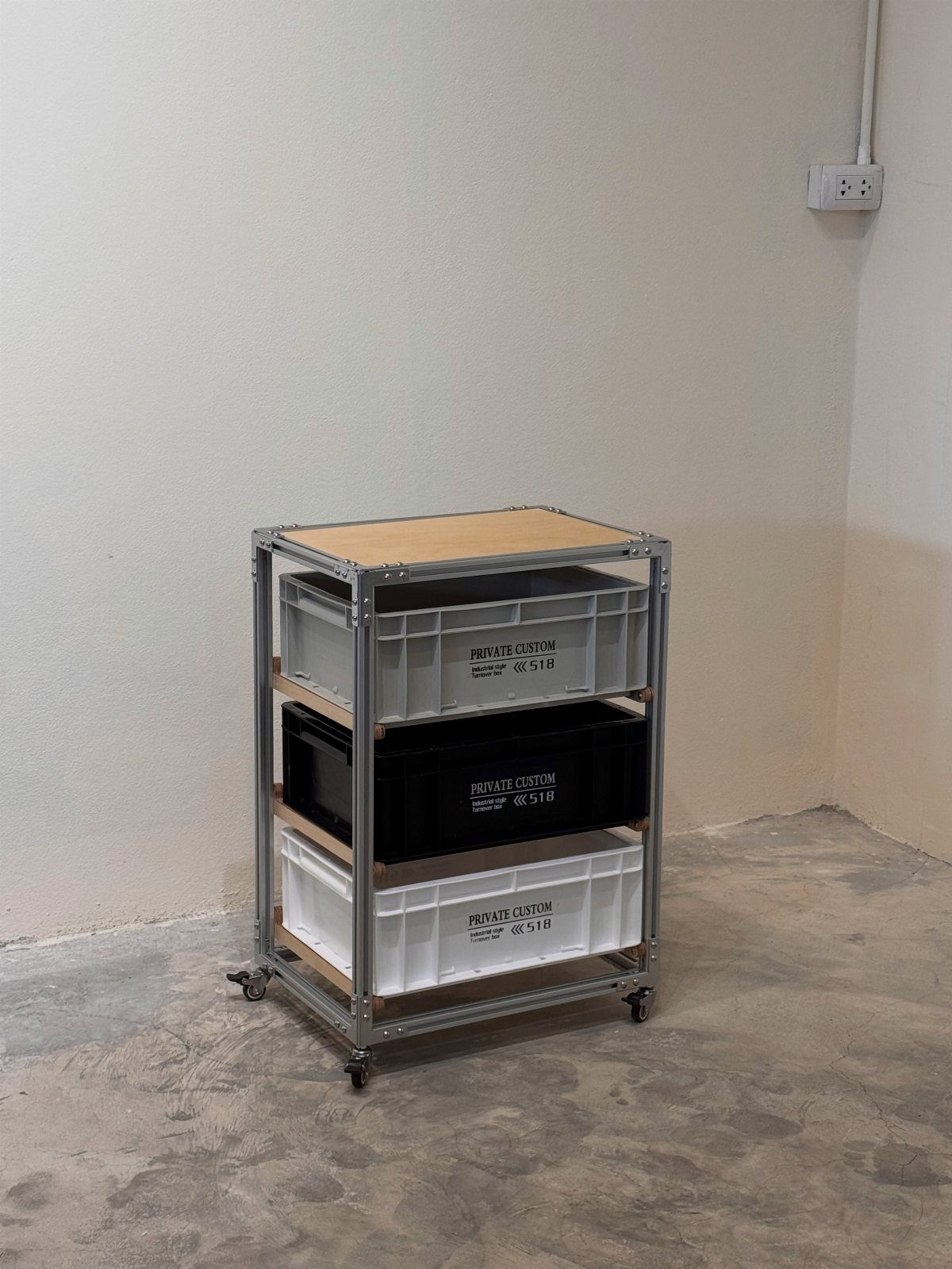 A metal rolling trolley with three large storage bins against a white wall.