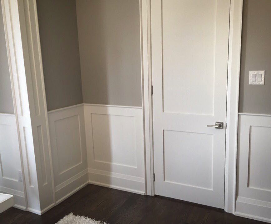 White flat panel wainscoting installed on grey walls next to a white door and dark floor.